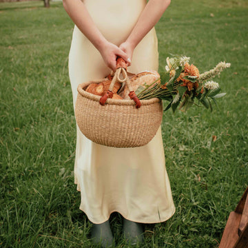 Woman in a yellow dress holding a basket and bouquet in a grassy field.