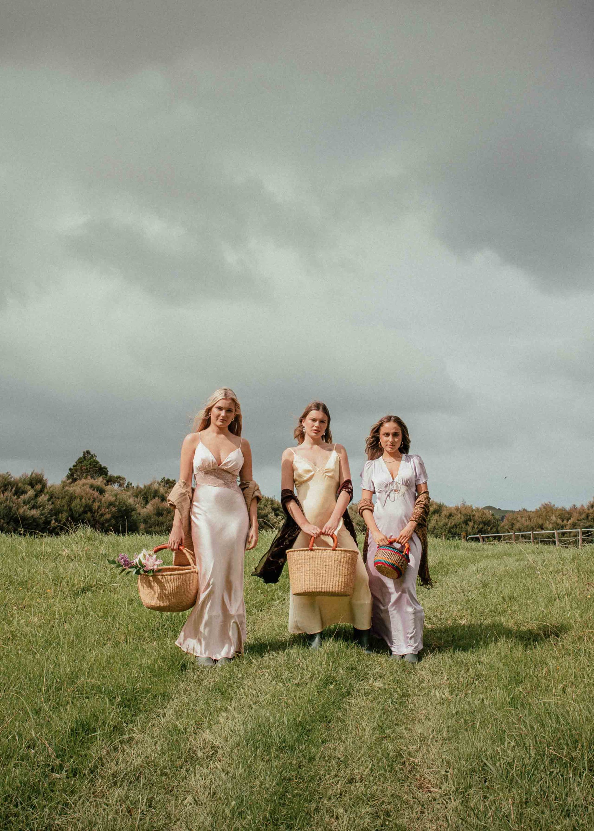 Three women walking together in a grassy field with a cloudy sky.