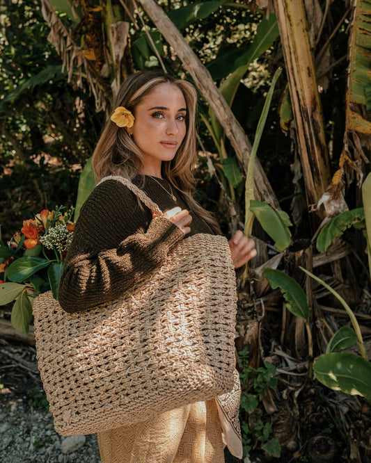 A woman wearing a dark brown knit sweater and beige skirt stands outdoors in front of tropical banana leaves, carrying a large woven straw tote bag.