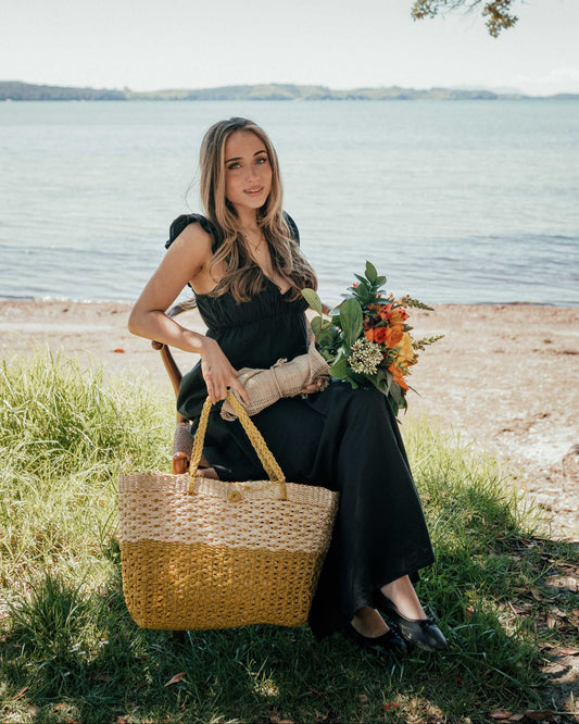 A woman in a black dress sits on a chair by the water holding a large woven straw tote bag and a colorful bouquet of flowers.