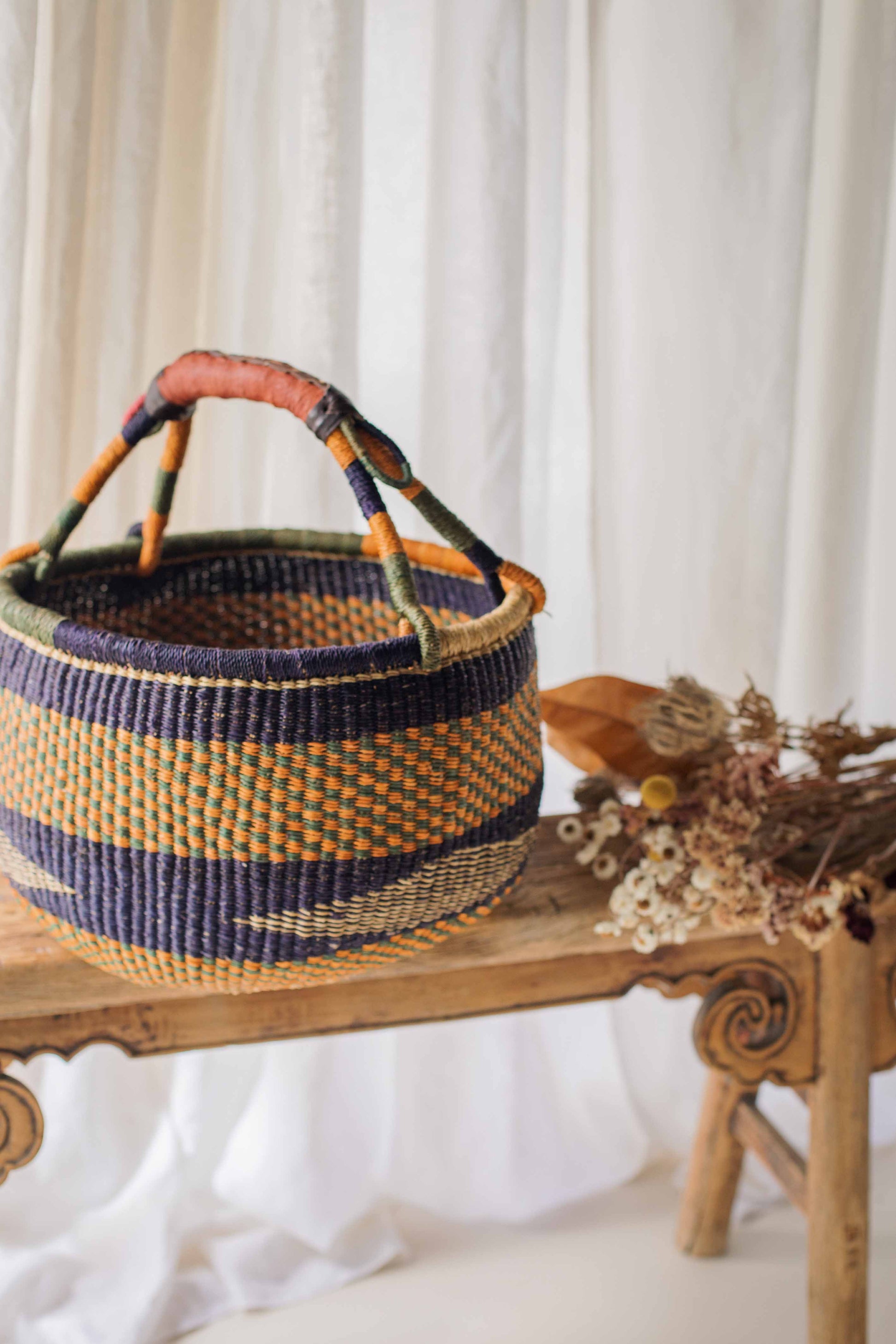Woven basket with colorful handles on a wooden surface with a white curtain background
