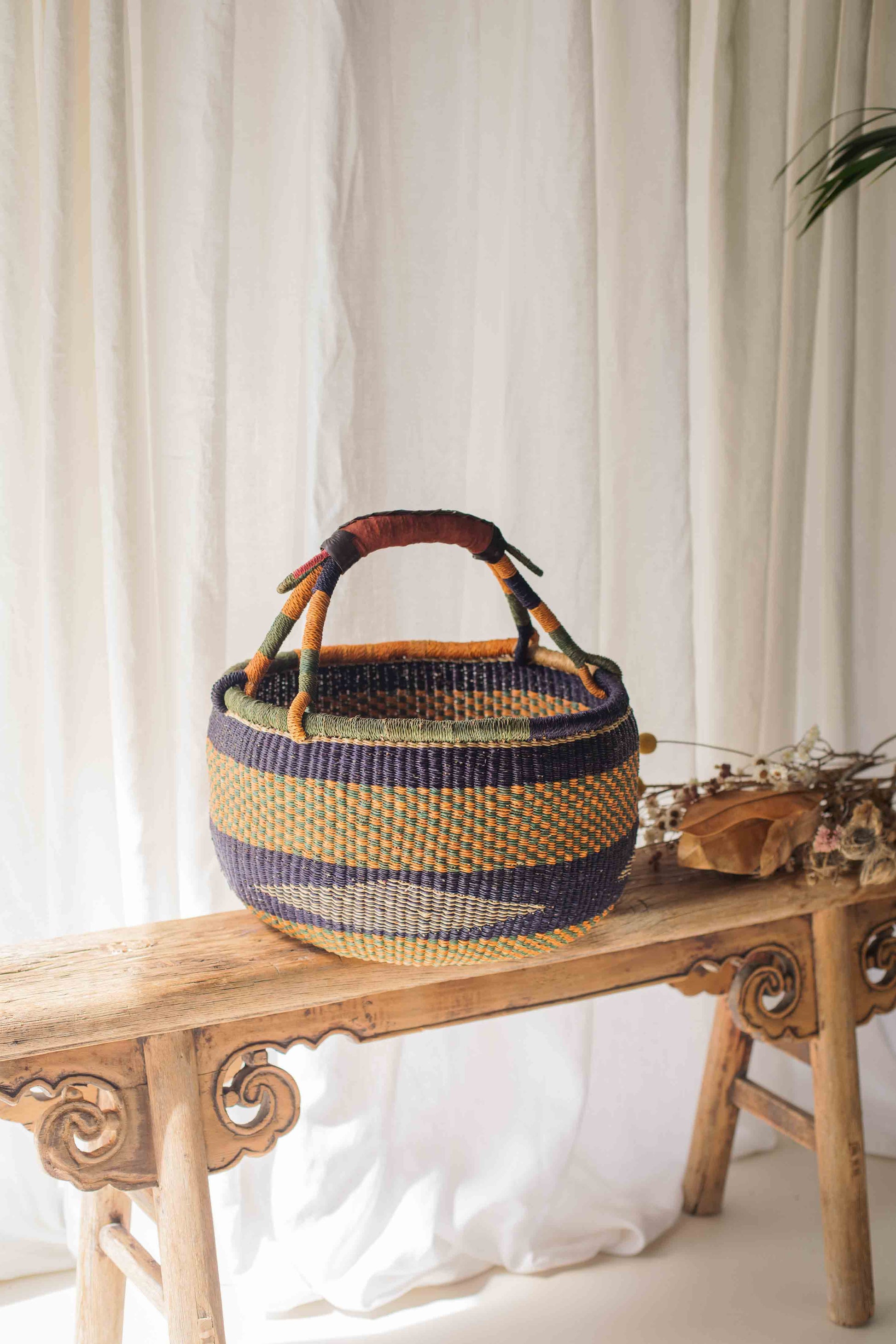 Woven basket with wooden handle on a wooden bench against a white curtain background