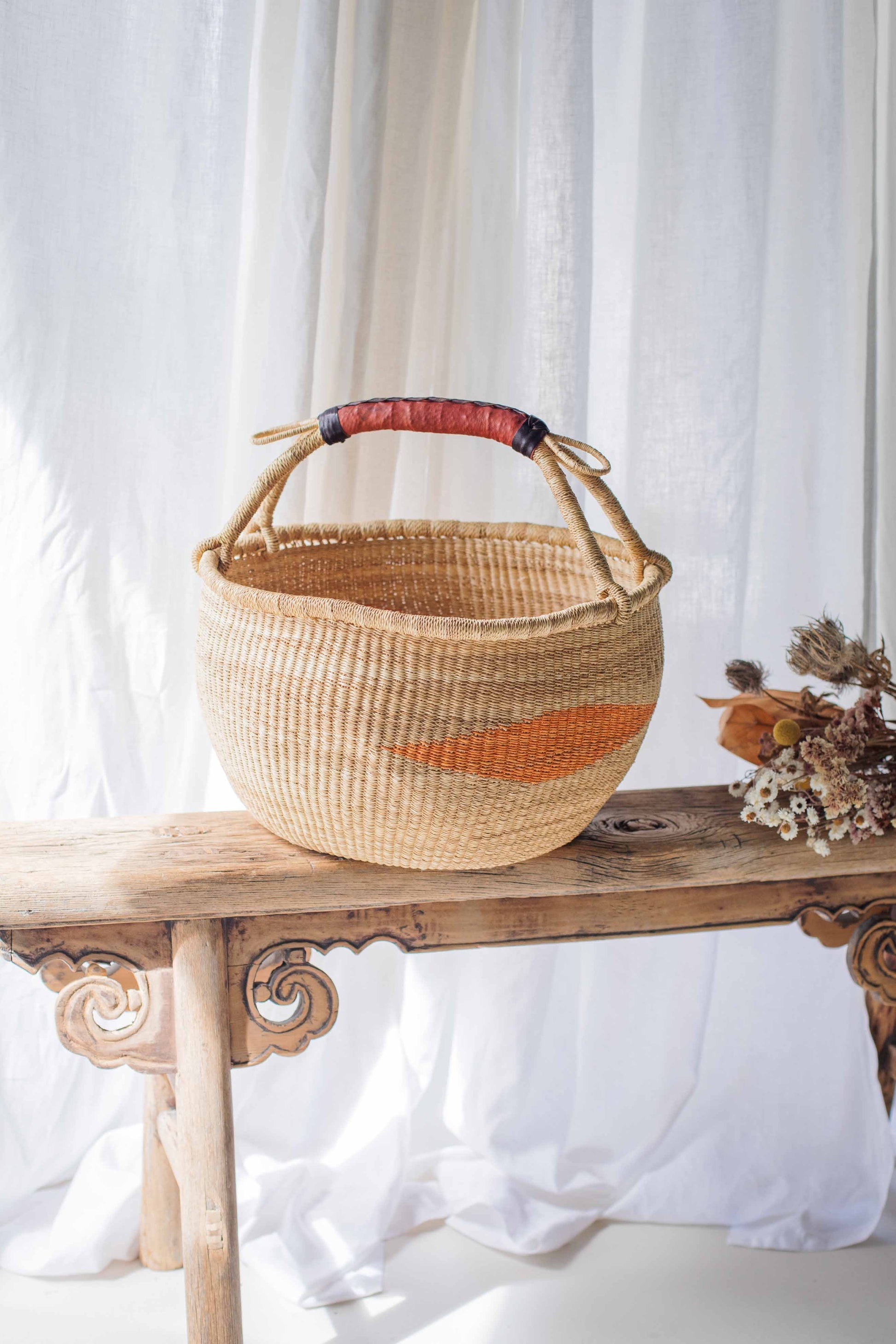 Woven basket with a red and orange stripe on a wooden bench against a white curtain background