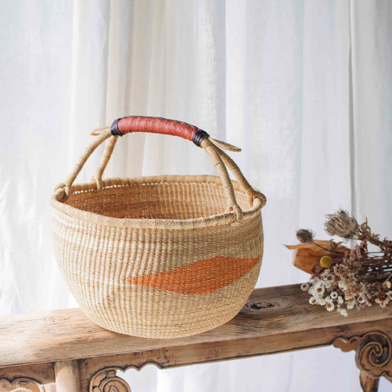 Woven basket with a red accent on a wooden bench against a white curtain background