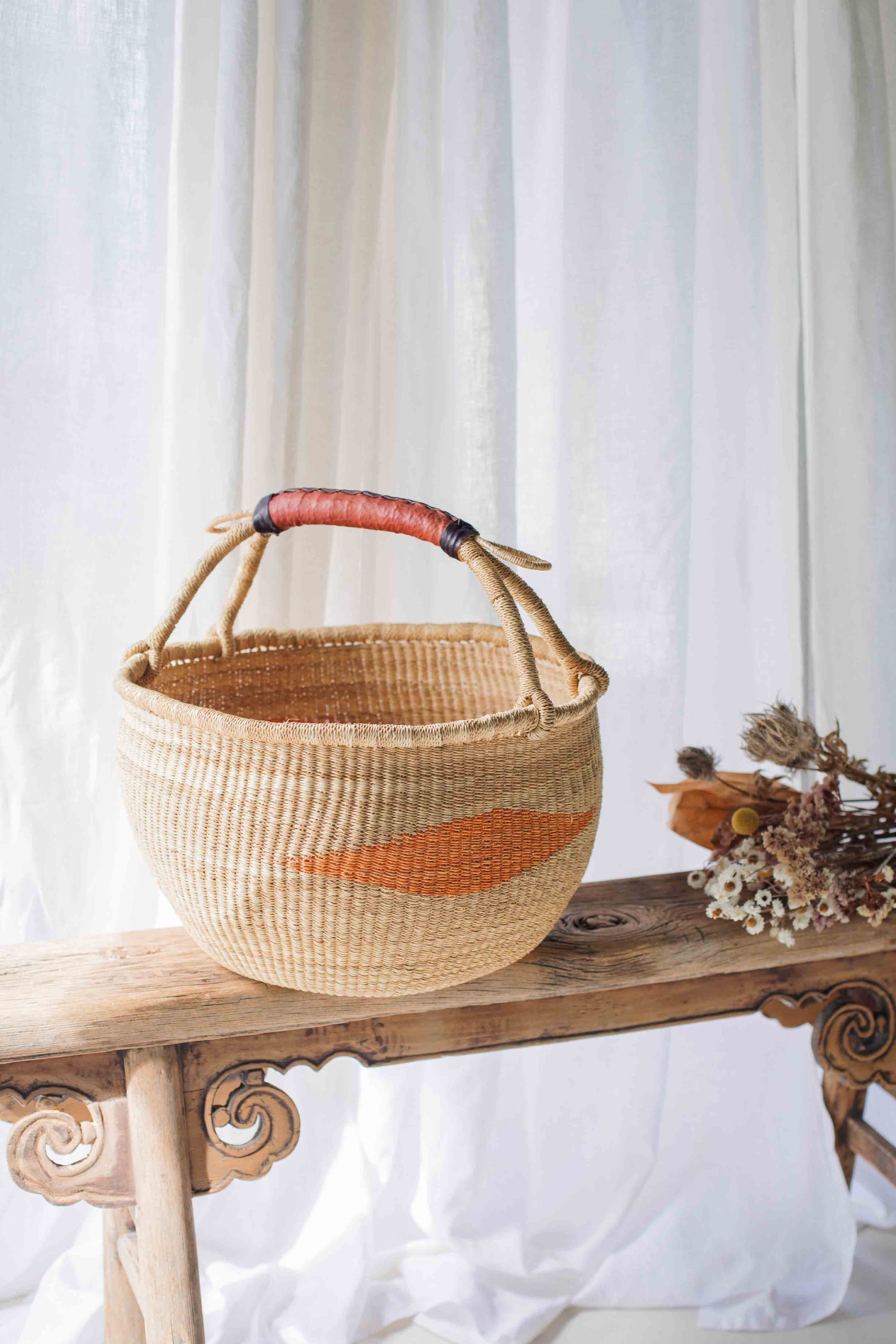 Woven basket with a red accent on a wooden bench against a white curtain background