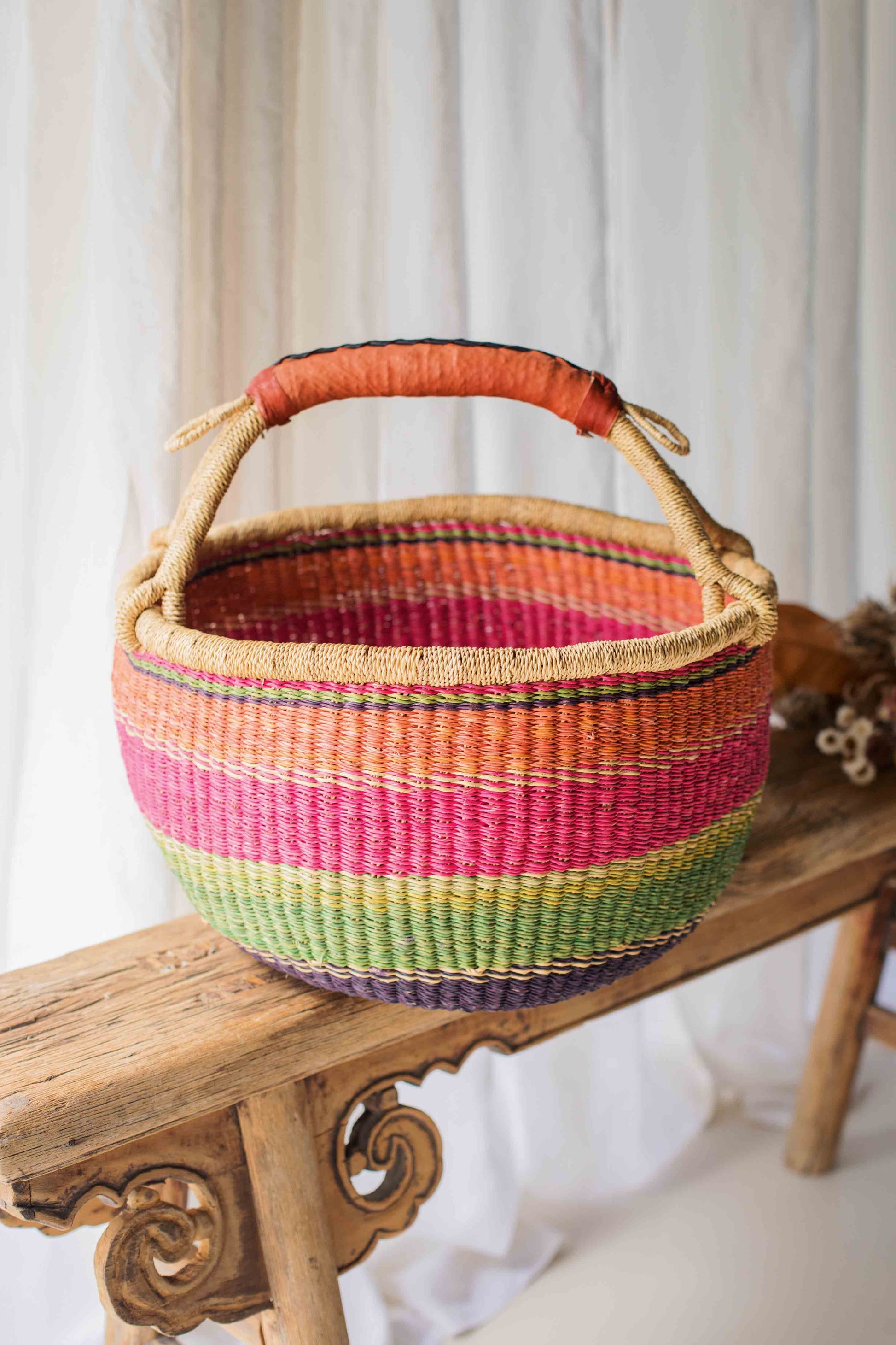 Colorful woven basket with a handle on a wooden bench against a white curtain background