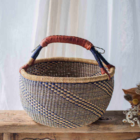 Woven basket with a brown handle on a wooden surface against a white curtain background