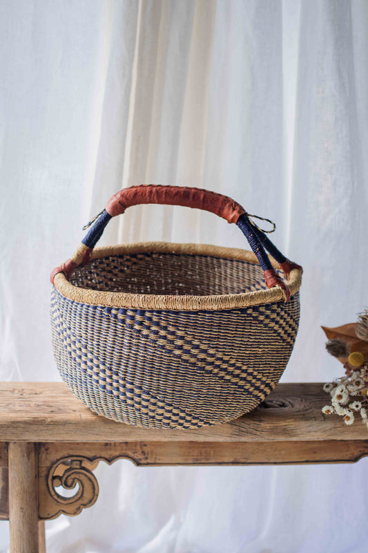 Woven basket with a brown handle on a wooden surface against a white curtain background