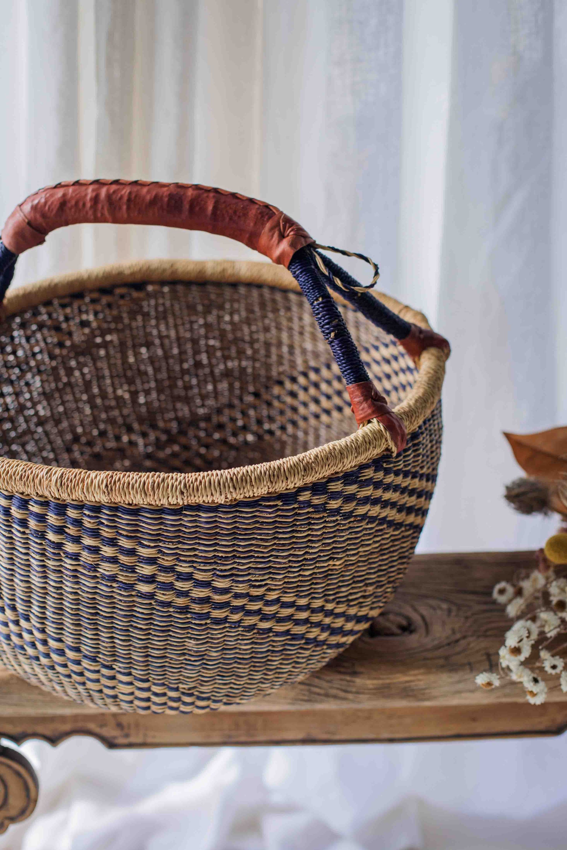 Woven basket with a red handle on a wooden surface with a white curtain background