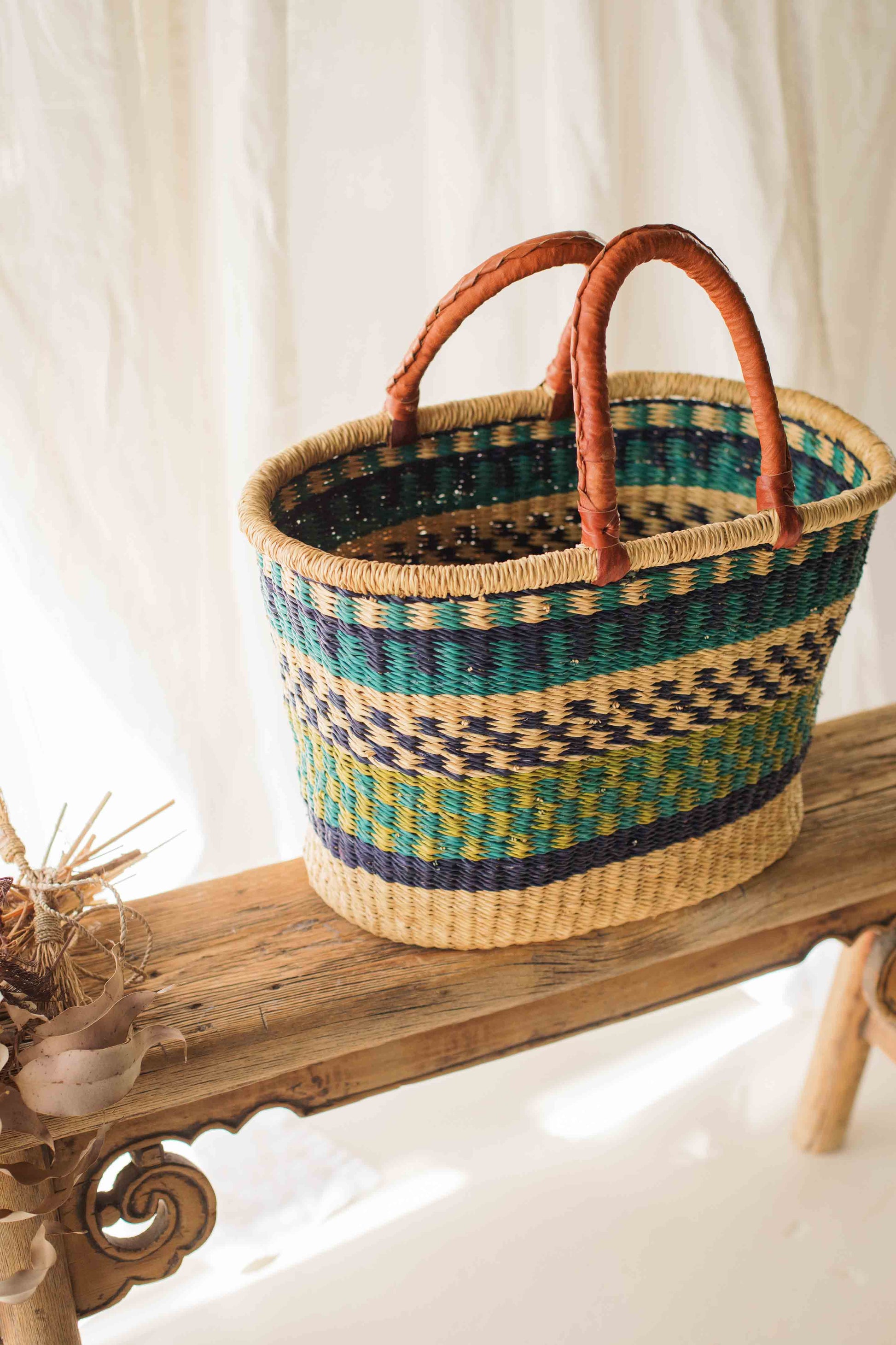 Woven basket with brown handles on a wooden surface against a white curtain background