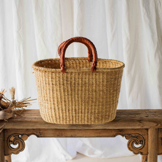 Woven basket with brown handles on a wooden table against a white curtain background