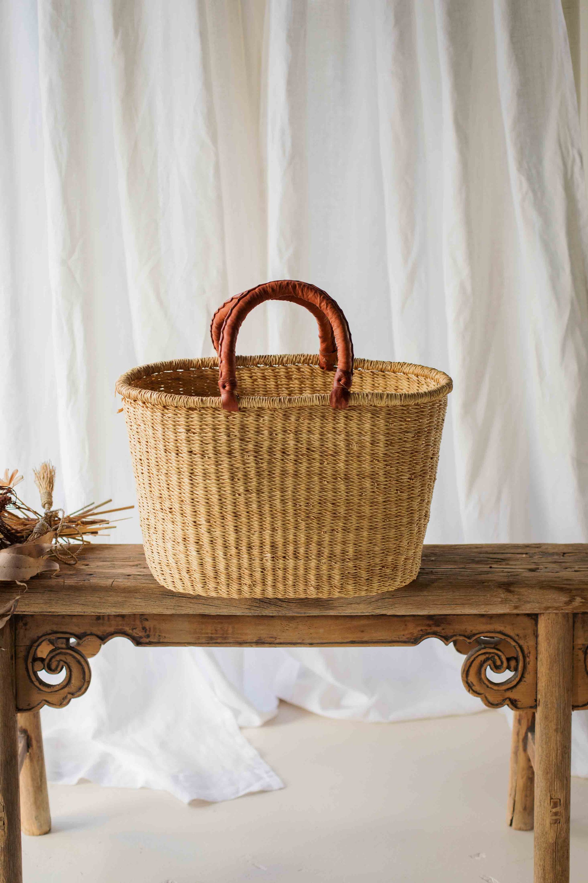 Woven basket with brown handles on a wooden table against a white curtain background