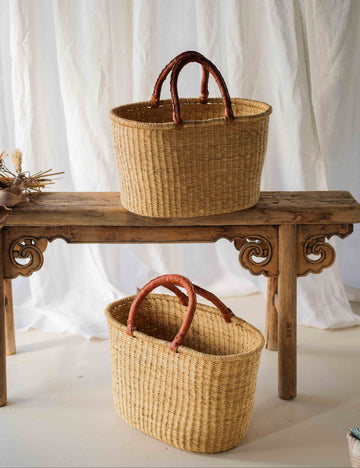 Two woven baskets with brown handles on a wooden table against a white curtain background.