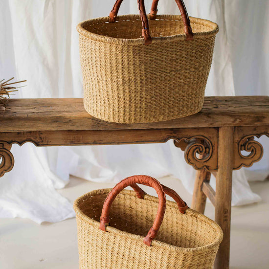 Two woven baskets with handles on a wooden table against a white curtain background.