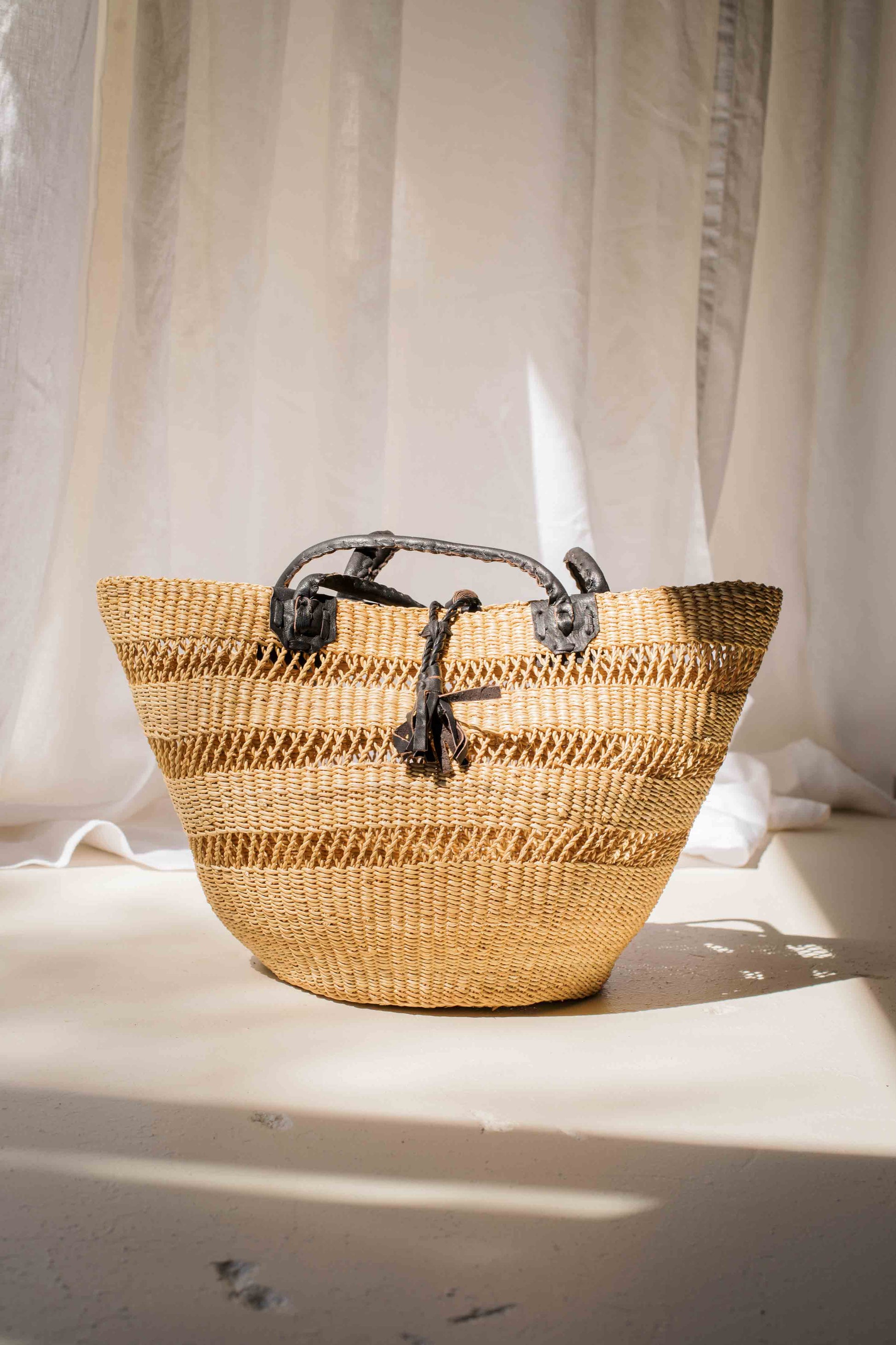 Woven basket with black handles on a light surface with white curtains in the background
