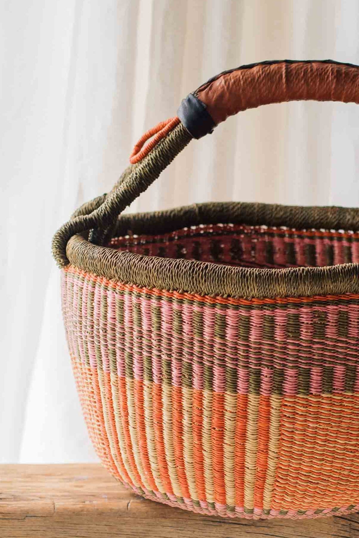 Woven basket with a brown handle on a wooden table against a white curtain background