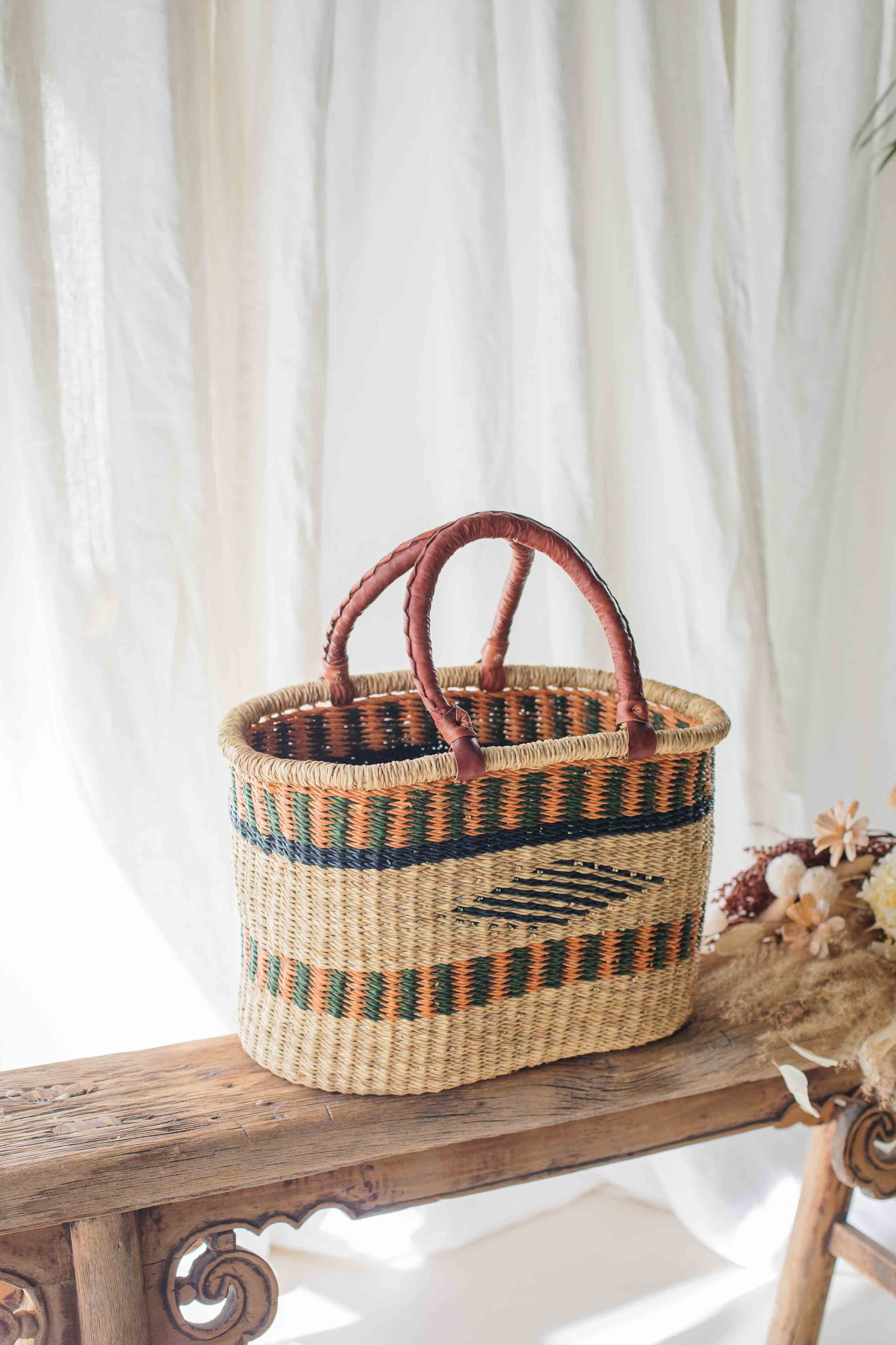 Woven basket with decorative patterns on a wooden bench against a white curtain background