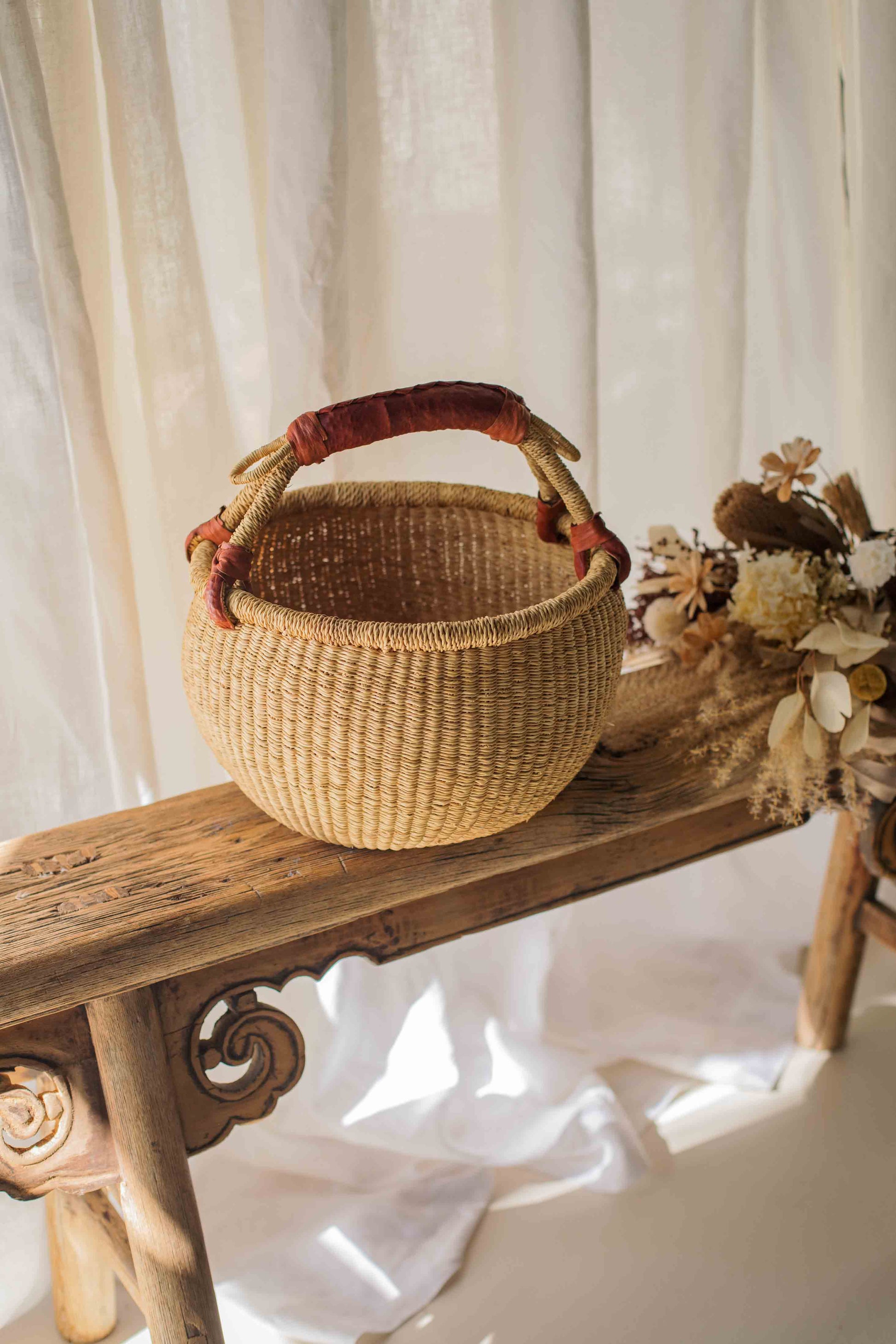 Woven basket with a red handle on a wooden bench against a white curtain background