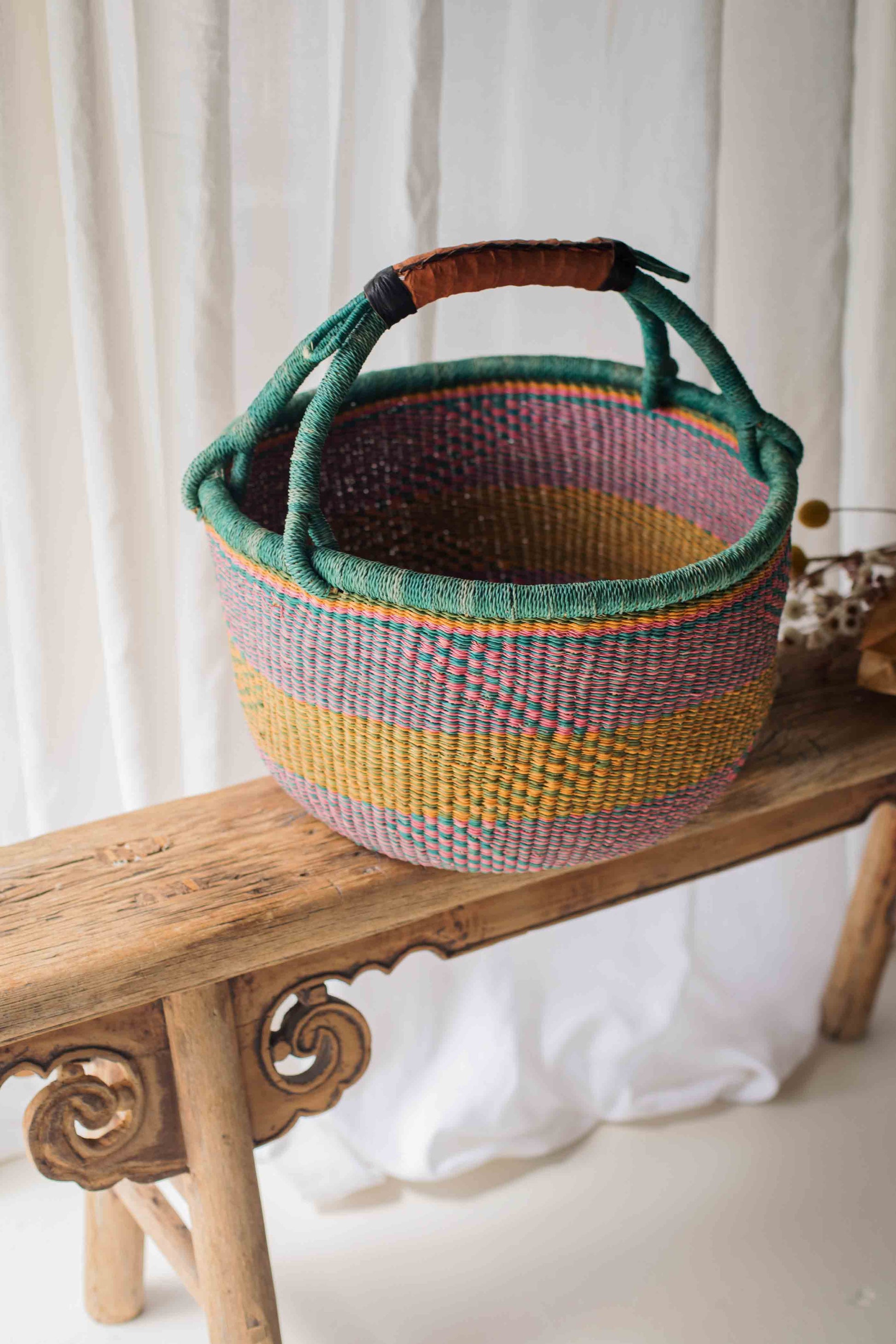 Colorful woven basket with a handle on a wooden bench against a white curtain background