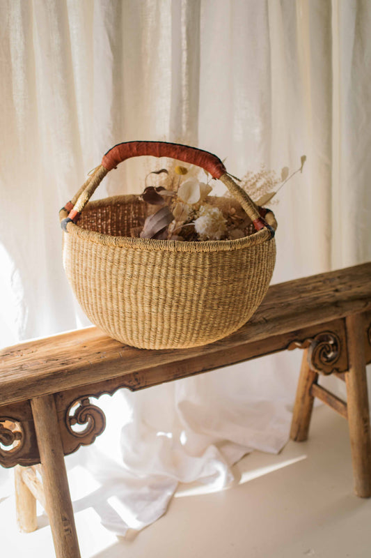 Woven basket with dried flowers on a wooden bench against a white curtain background