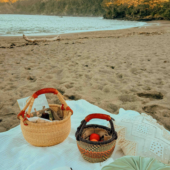 A beach picnic setup on a white blanket with a woven basket, a small dark basket with red handles, an open book, and a green cushion, with a sandy beach and ocean in the background.