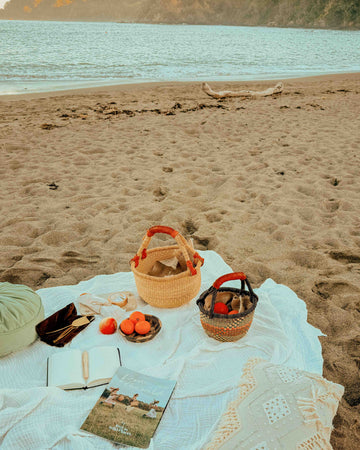 A styled beach picnic setup on a white blanket on sandy shore, with baskets, fruit, a book, sunglasses, and a pillow, overlooking a calm body of water and distant hills.