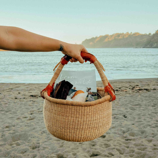 A woven straw beach tote bag with orange-red handles is held by a person's arm on a sandy beach with the ocean and hills in the background.