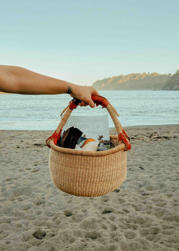 A woven straw beach tote bag with orange-red handles is held by a person's arm on a sandy beach with the ocean and hills in the background.
